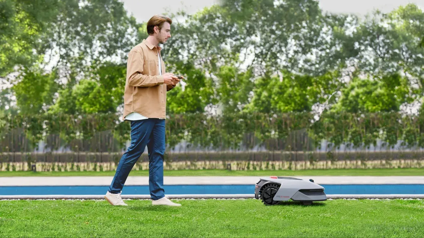 Un homme consulte son téléphone pendant que le Dreame A1 Pro tond une pelouse au bord d'une piscine